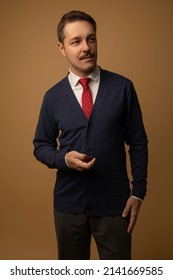 Studio Portraits Of A Man Dressed In 1960s Clothing. He Is Wearing A Cardigan With A Button Up Shirt Underneath And A Red Tie. He Is Standing Up Holding A Pipe. The Background Is Beige.