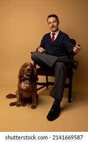 Studio Portraits Of A Man Dressed In 1960s Clothing. He Is Wearing A Cardigan With A Button Up Shirt Underneath And A Red Tie. He Is Sitting In A Chair And A Cocker Spaniel Dog Is Sitting Beside Him.