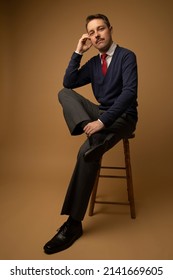 Studio Portraits Of A Man Dressed In 1960s Clothing. He Is Wearing A Cardigan With A Button Up Shirt Underneath And A Red Tie. He Is Sitting On A Wooden Stool. The Background Is Beige. 