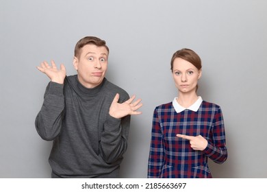 Studio Shot Of Adult Couple, Serious Woman Is Pointing With Index Finger At Man Making Goofy Face, Having Fun And Fooling Around, Both Are Standing Over Gray Background