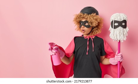 Studio Shot Of Amazed Curly Haired Woman Dressed In Black T Shirt Eyemask Rubber Gloves And Cape Uses Cleaning Detergent For Tidying Up Holds Mop Going To Wash Floor Isolated Over Pink Background