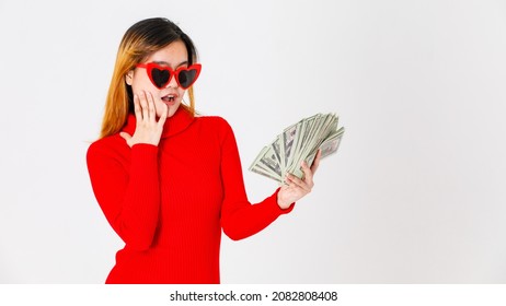 Studio shot of Asian lucky female fashion model in casual fashionable trendy red dress with heart shape sunglasses win lottery prize holding dollar cash banknote currency fan on white background.