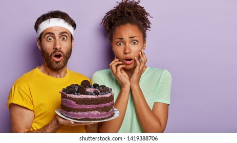 Studio Shot Of Astonished Mixed Race Couple Stare With Disbelief, Hold Plate With Delicious Fruit Cake, Celebrate Birthday, Stand Over Purple Background. Dessert, Fast Food And Dieting Concept