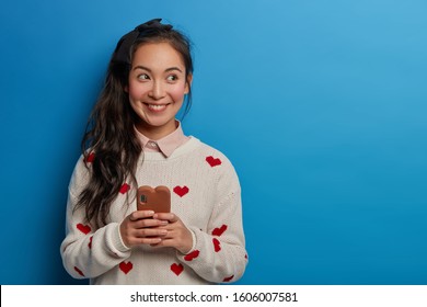 Studio Shot Of Atrractive Asian Woman Has Pony Tail, Dressed In Casual Jumper, Looks Aside, Wears Jumper, Surfs Internet On Mobile Phone, Poses Against Blue Wall. Happy Emotions, Technology.