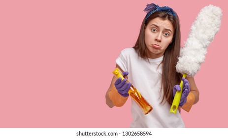 Studio Shot Of Attractive Woman Purses Lower Lip, Dressed In Casual Clothes, Holds Dust Brush And Cleaning Detergent, Models Against Pink Background With Blank Space For Your Promotional Content