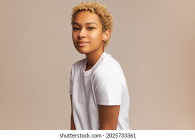 Studio shot of attractive young lady of mixed-race posing with calm, peaceful face expression, gently smiling at camera, having trendy, natural look, wearing nose ring and short blonde haircut