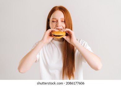 Studio Shot Of Attractive Young Woman With Closed Eyes Enjoying Bite Of Appetizing Delicious Hamburger On White Isolated Background. Closeup Front View Of Female Eating Tasty Burger .