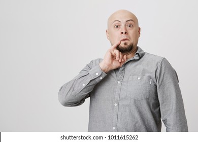 Studio shot of bald bearded man in gray shirt poses against gray background, holds little finger behind corner of his mouth, looks like Dr. Evil. Hairless male grimacing and making faces indoors