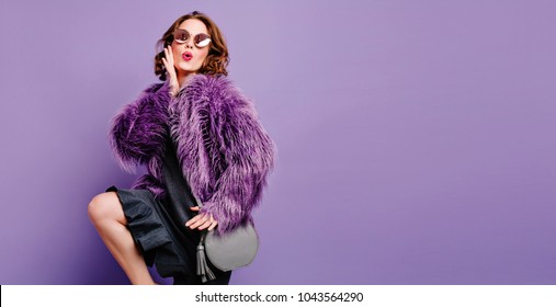 Studio shot of barefooted cute woman with trendy gray purse posing with pleasure. Place for text. Indoor portrait of relaxed girl in sunglasses dancing on purple background.