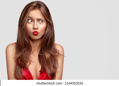 Studio shot of beautiful brunette woman makes grimace, pouts lips, raises eyebrows, has healthy clean skin, wears red dress, poses against white background with blank space for your advertisement