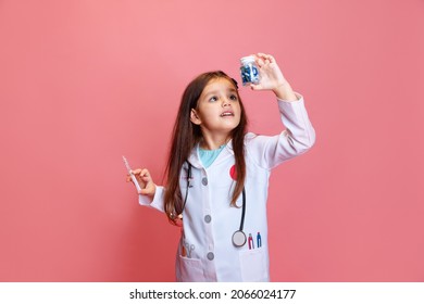 Studio Shot Of Beautiful Little Girl, Child Playing Doctor Wearing White Lab Coat Posing Isolated On Pink Studio Background. Concept Of Childhood, Studying, Leisure Games. Health Care And Medicine.