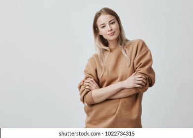 Studio Shot Of Beautiful Young Woman With Blonde Straight Hair Smiling Gently While Listening To Interesting Conversation, Wearing Loose Long-sleeved Sweater, Keeping Arms Folded. Beauty And Youth