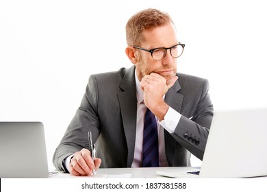 Studio shot of a businessman wearing suit and looking thoughtfully while sitting at desk and working on laptop. Isolated on white background. - Powered by Shutterstock