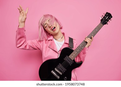 Studio shot of carefree female musician performs favorite music keeps arm raised has pink hair floating on wind holds black acoustic guitar enjoys rock n roll dreams to become famous artist. - Shutterstock ID 1958073748