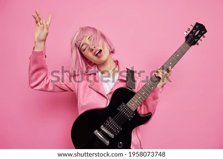 Studio shot of carefree female musician performs favorite music keeps arm raised has pink hair floating on wind holds black acoustic guitar enjoys rock n roll dreams to become famous artist.