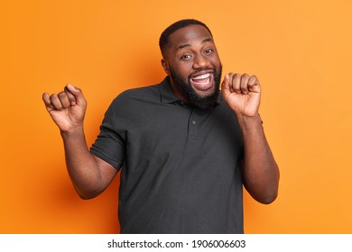 Studio shot of carefree positive black man with thick beard dances carefree has upbeat mood raises arms enjoys favorite music isolated over vivid orange wall. Afro American guy moves actively indoor - Shutterstock ID 1906006603