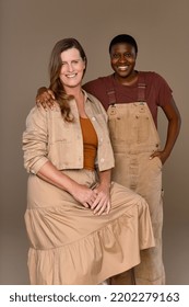 Studio Shot Of A Caucasian Transgender Woman In Her 50's And An Afro-Latino Person Their 20's Smiling Together On A Neutral Background.