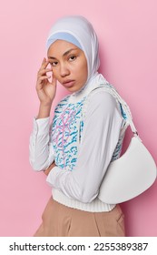 Studio shot of charming beautiful Arabic woman touches face gently has healthy skin wears hijab white shirt and vest poses with bag on shoulder isolated over pink background. Religious female model - Shutterstock ID 2255389387