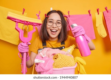 Studio Shot Of Cheerful Asian Woman Looks Happily At Camera Enjoys Finishing Housework Holds Cleaning Detergent And Mop Does Housekeeping Poses Over Rope With Drying Clothes. Daywork Routine