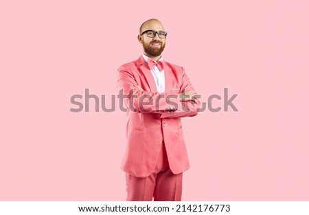 Studio shot of cheerful young man in pink suit. Happy confident bald guy with ginger beard, wearing pink suit, bow tie and stylish glasses standing arms folded isolated on solid pink color background