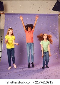 Studio Shot Of Children With Glitter Jumping In The Air With Outstretched Arms On Purple Background