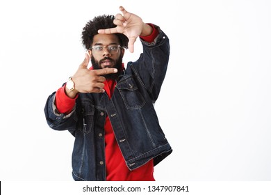 Studio Shot Of Cool And Stylish African American Male Producer In Glasses Stepping Back As Pulling Hands Towards Camera With Frames Gesture Looking Through, Picturing Scene From Clip Over Gray Wall