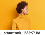 Studio shot of curly woman stands sideways against yellow background turns head aside has serious expression dark curly hair dressed in turtleneck wears round earrings notices something on right