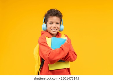 Studio shot of a cute smiling African American kid student wearing orange hoodie, bright backpack and wireless headphones, posing over yellow background with a pile of books  in hands. - Powered by Shutterstock