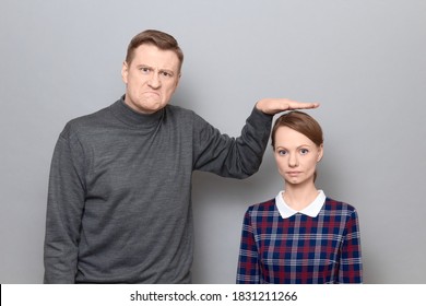 Studio shot of disgruntled tall man showing height of short woman, both are wearing casual clothes, standing over gray background. Concept of diversity of people's heights, tall and short persons