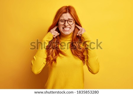 Studio shot of dissatisfied redhead woman clenches teeth and plugs ears annoyed by loud sound or noise wears casual jumper isolated on yellow background. Female complains on music from outside
