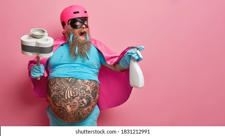 Studio Shot Of Emotional Cleaning Man Superhero Shouts Loudly And Looks Away Poses With Detergent Plunger Stands Against Pink Background With Copy Space For Your Promotional Content. Day Of Washing