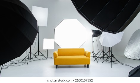 Studio shot fashion backstage photographing shooting set with yellow cozy sofa couch and photography equipment softbox flash strobe spotlight tripods reflector umbrella on white backdrop background. - Shutterstock ID 2100157552