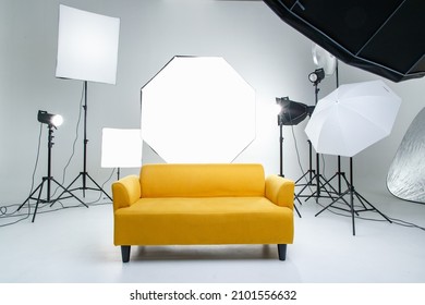 Studio shot fashion backstage photographing shooting set with yellow cozy sofa couch and photography equipment softbox flash strobe spotlight tripods reflector umbrella on white backdrop background.