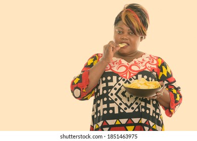 Studio shot of fat black African woman eating and holding bowl of potato chips