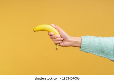 Studio Shot Of Female Hand Holding A Banana Like A Gun Isolated On Bright Colored Orange Yellow Background. Joke, Humor, Comedy Concept.