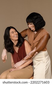 Studio Shot Of A Filipino Woman In Her 50's And An African American Woman With Vitiligo In Her 30's Looking At Each Other And Smiling On A Neutral Background.