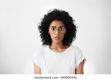 Studio shot of frightened bug-eyed young female feeling scared and shocked, watching horror movie. Attractive dark-skinned woman dressed casually having terrified look while receiving shocking news