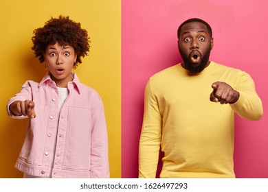 Studio Shot Of Frightened Emotive Ethnic Couple Point Index Fingers At Camera, See Surprising Thing In Front, Gasp From Fright, Keeps Eyes Widely Opened, Isolated On Yellow And Pink Background.