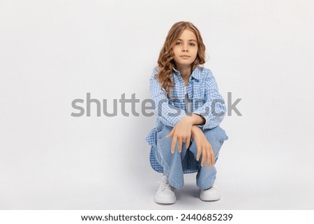 Studio shot front view young girl with blonde curly hair in casual clothes squatting on white background. Copy space. Teenager concept.