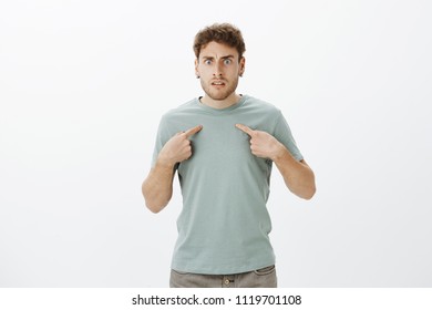 Studio shot of frustrated angry young man with bristle in t-shirt and earrings, pointing at himself and frowning, being insulted or offended by friend, standing outraged over gray wall