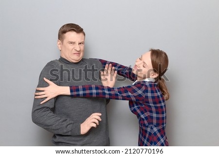 Studio shot of funny couple, happy cheerful woman is trying to hug man resisting and grimacing from displeasure, both are standing over gray background. Relationship concept, human emotions