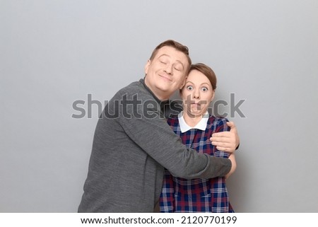 Studio shot of funny couple, happy cheerful man is hugging shocked woman expressing bewilderment, looking stunned, both are standing over gray background. Relationship concept, human emotions