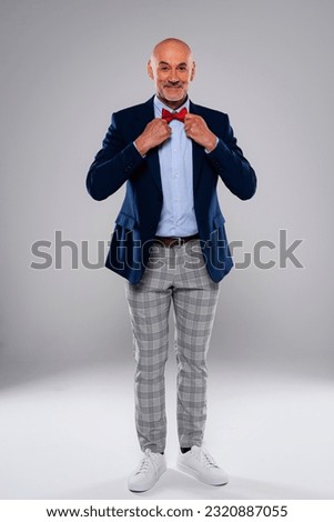 Studio shot of funny faced man checked pants and bow tie while standing at isolated grey background. He is adjusting his bow tie. Copy space.