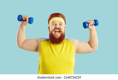 Studio shot of funny happy excited bearded redhead plump fat man in yellow sweatband and sleeveless top doing sports exercise with dumbbells, showing his strength and smiling. Training workout concept