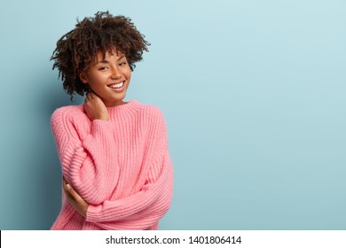 Studio shot of glad charming young female with Afro haircut, touches neck, wears oversized jumper, isolated over blue background with blank space for your promotional content. Pleasant emotions