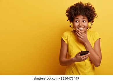 Studio shot of glad dark skinned curly teenager has fun indoor, listens pleasant melody in headphones, looks happily aside, wears bright yellow t shirt, feels relaxed. Monochrome. Hobby concept - Powered by Shutterstock