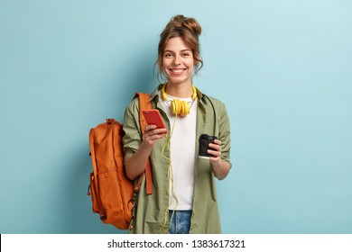 Studio shot of glad female student has coffee break after lectures, listens audio book in headphones, enjoys record from website, uses mobile phone for chatting online, has rucksack on back. - Powered by Shutterstock