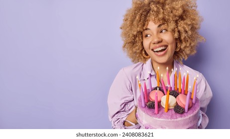 Studio shot of glad woman with curly bushy hair holds big tasty cake looks aside happily smiles broadly wears festive clothing celebrates birthday isolated over purple background copy space for text - Powered by Shutterstock