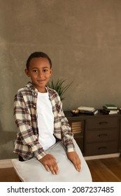 Studio Shot Of God-looking Shy Boy Standing Against Grey Wall And Vintage Book Case, Looking At Camera, Putting Hands On Couch Pillow. Positive Human Emotions. Children, Kids, Childhood Concept