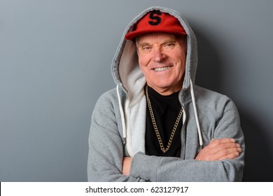 A Studio Shot With A Gray Wall Behind A Man. Funny Grandad Gives A Smile. A Red Cap, A Gray Hoodie And A Black T-shirt With A Gold Chain On It Make This Grandpa Look Younger.